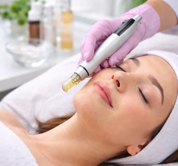 Licensed esthetician using a nano infusion device on a client&rsquo;s cheek in a clean professional treatment room, with serum visible on the skin surface.